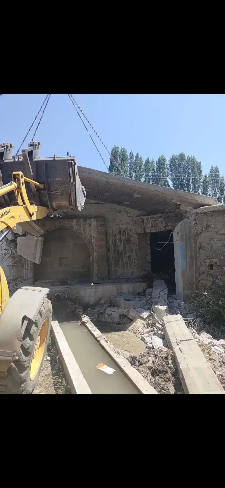 Taş duvarlı yapının betonarme saçağını kovayla zorlayan sarı lastikli yükleyici ve yapının önünden geçen sulama kanalı